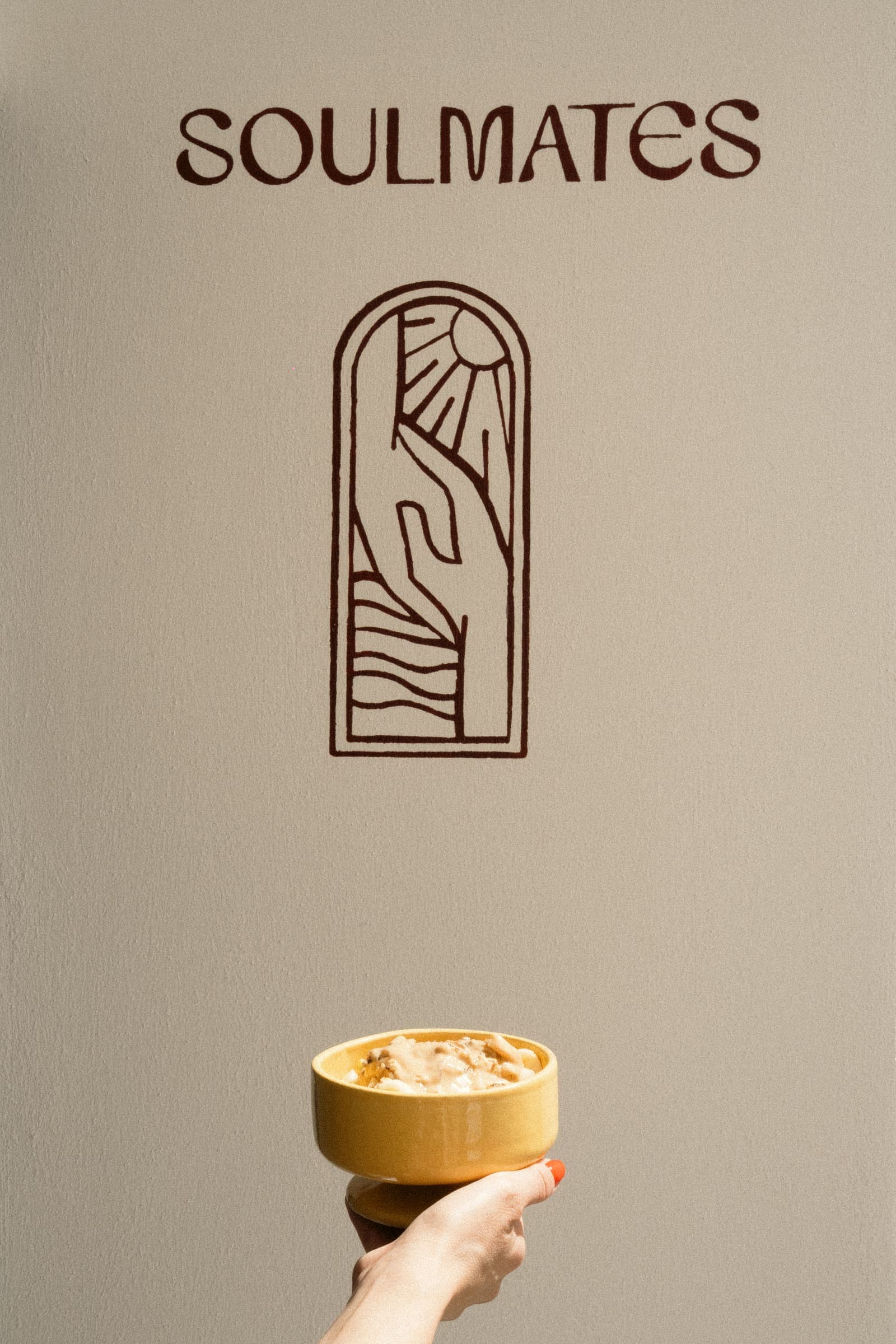 A hand lifts a yellow bowl of creamy porridge toward the Soulmates wordmark and arched hand-and-sunburst logo painted in dark brown on a warm beige wall.